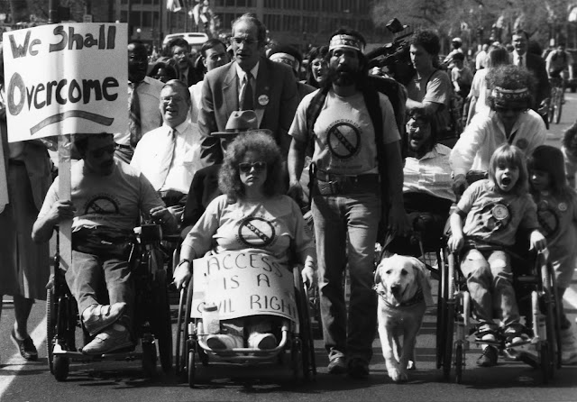 A black and white photo of the “Wheels of Justice” March in Washington, D.C. on March 12, 1990. There are people with disabilities who use wheelchairs holding signs that say, "We Shall Overcome" and "Access is a Civil Right." There is a blind man walking with his guide dog and a child with a disability being pushed in a wheelchair. 