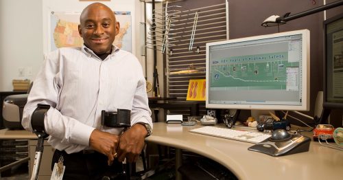 Man sitting at a desk holding forearm crutches and working at a computer at the national highway transportation administration.
