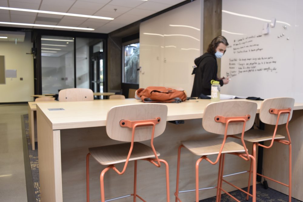 Student using the white board while their notes and backpack rest on a wooden table surrounded by chairs