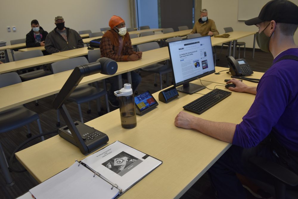 Students attending a class, photo highlights the instructor using the height adjustable table for their lecture with touch panel, computer and document projector