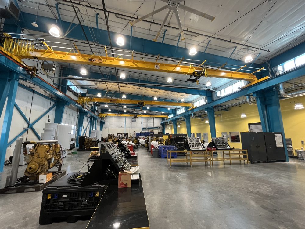 Interior view of the high bay area in the Dealer Service Tech Building showing large engines in the bay, roll up doors on the left, and cranes on steel beams on the ceiling