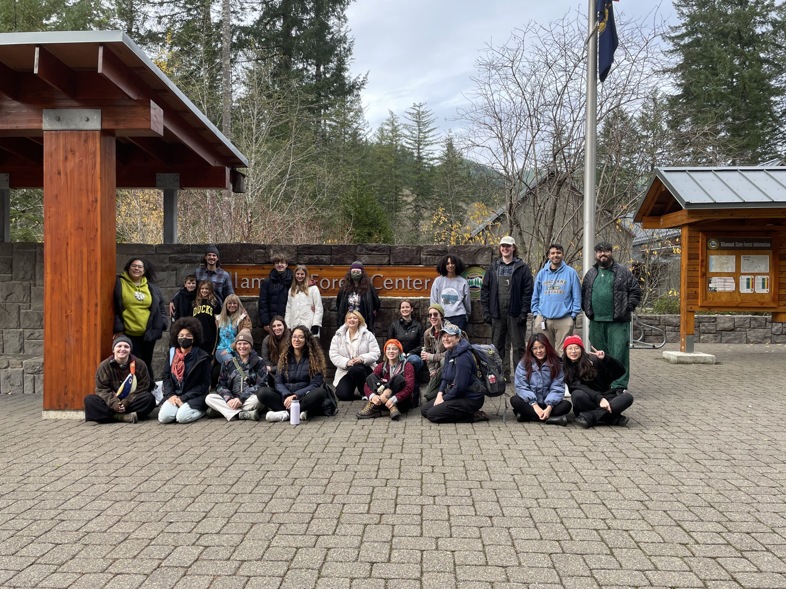 A group of PCC students, staff, and friends and family members are grouped in front of a wooden sign that says 'Tillamook Forest Center'. Some are sitting, others are standing, all have smiles on their faces as they look at the camera. 
