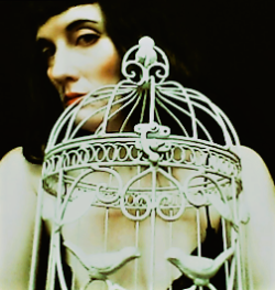 A moody photo of a person behind a small ornate white birdcage looking at the camera.