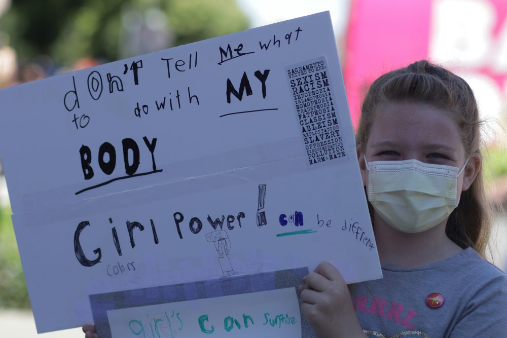 A masked young girl holding a hand-drawn sign that reads “don’t tell me what to do with my body! Girl power can be different colors”.