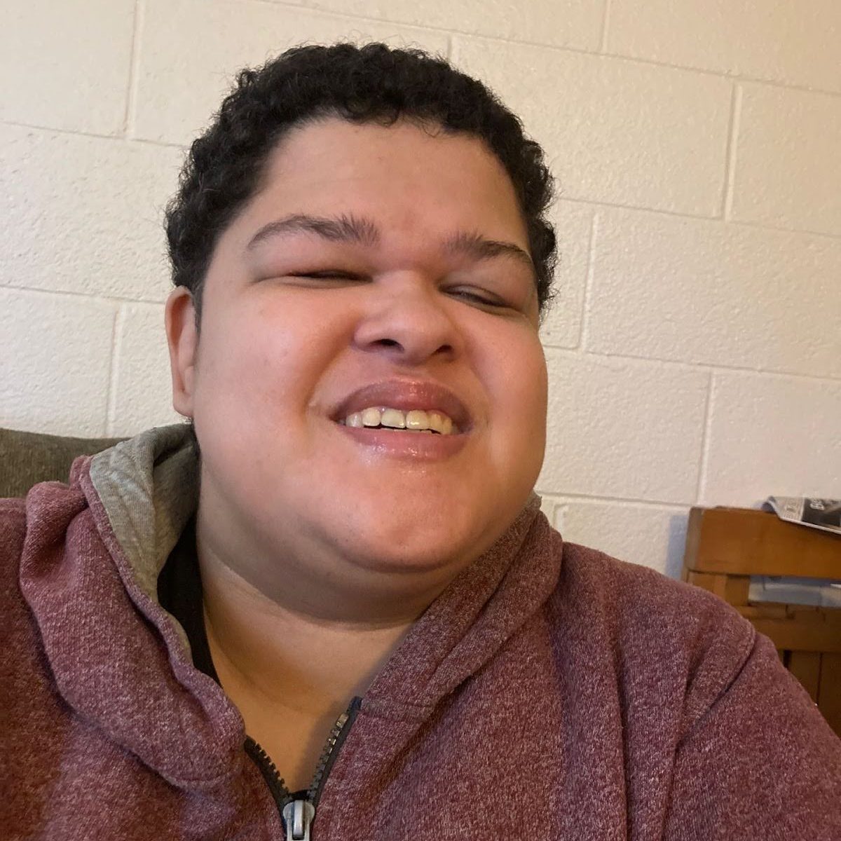 A half half body profile photo of podcast tech Cherranne, a blind woman with medium skin tone and short dark curly hair, wearing a zip up hoodie and smiling at the camera.