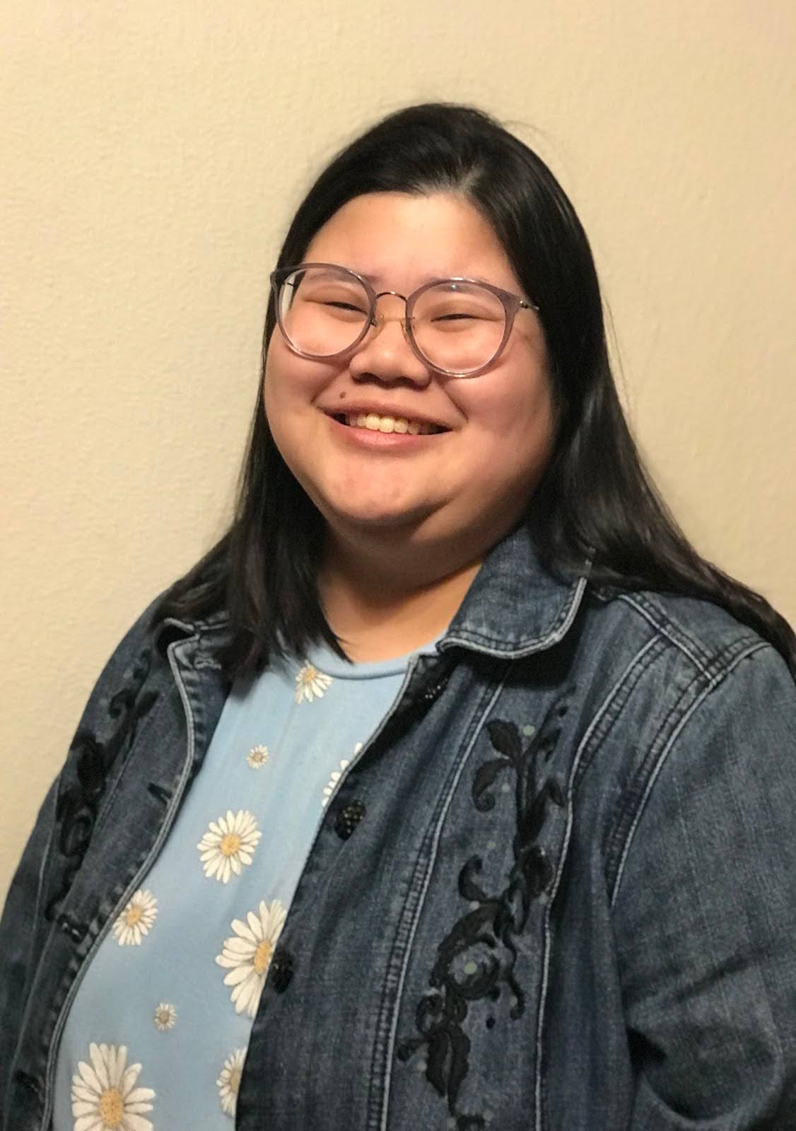 Co-lead producer, Asher. A half body profile image of a young Asian woman wearing glasses, Asher, stands in front of a blank backdrop, smiling at the camera. She wears a light blue blouse with white daisies of different sizes all over the blouse and a dark blue jean jacket with black plant embellishments on the chest.