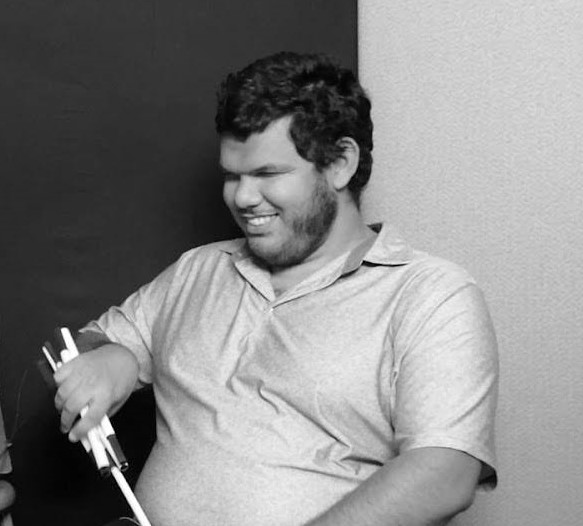 Black-and-white photo of podcast tech Miguel, a blind man with medium-light skin tone and short darkly colored hair wearing a polo shirt folds up his white-cane while sitting down to an interview for Let’s Talk! 