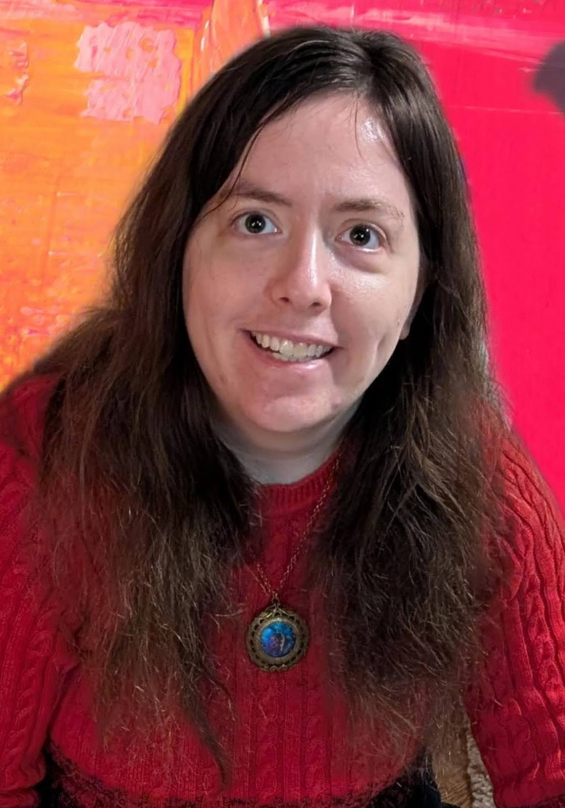 Half body profile photo of co-lead producer Amanda, a person with light skin tone, brown eyes, and long dark brown hair, smiling and wearing a red sweater and a gold and blue pendant necklace.