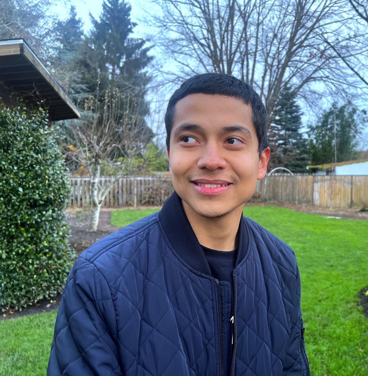 Half body profile photo of podcast tech Ricardo Bravo, a person with medium skin tone, short black hair, wearing a navy blue jacket and smiling while looking to the side. A grassy lawn, trees, bushes, and a fence are visible in the background.