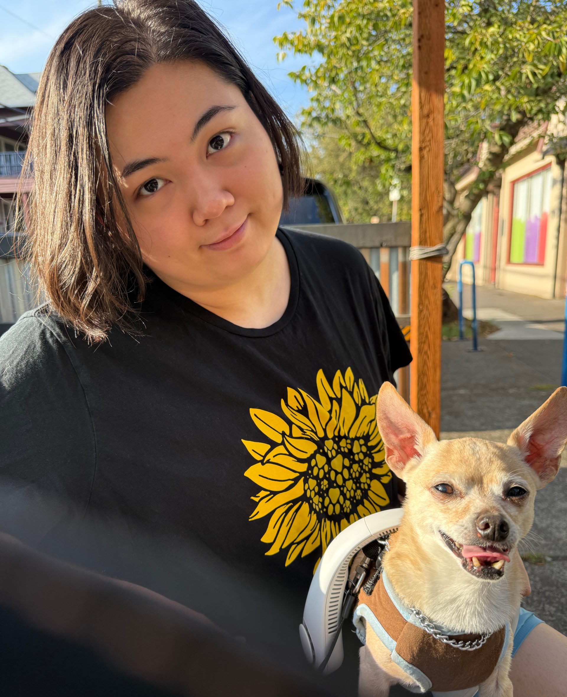 Podcast tech Julie, a person with light-medium skin tone, and shoulder-length dark brown hair, is smiling at the camera. Julie is sitting outdoors with a small dog with off-white and tan-colored fur, named Tako, sitting on their lap. Julie is wearing a black shirt with a yellow sunflower image on it.