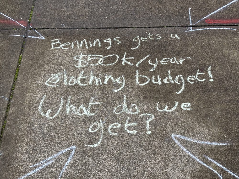 Chalk art on sidewalk that reads: "Bennings gets a $50k/ year clothing budget! What do we get?"