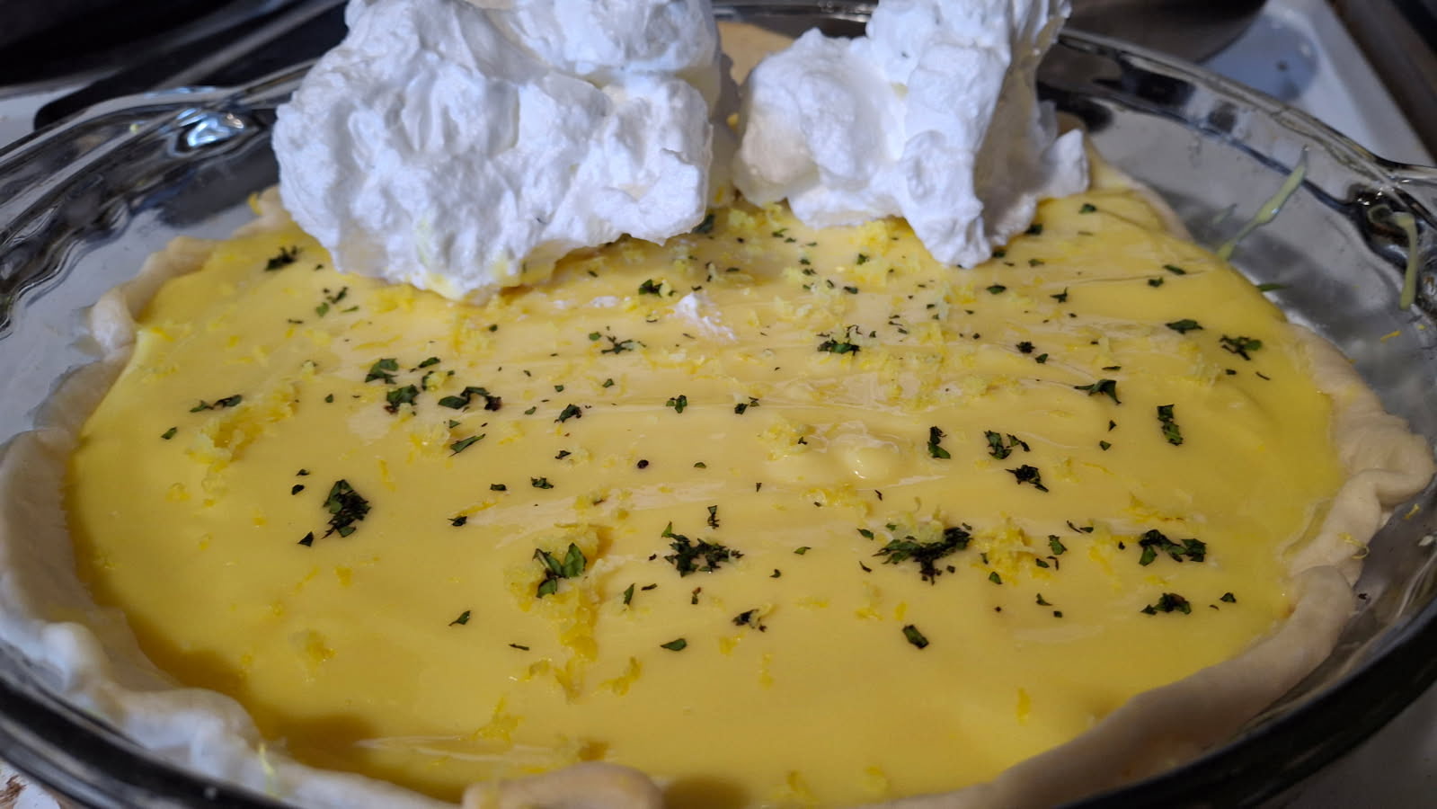 A close-up of a homemade Lemon Dream “Ice Box” pie in a glass pie dish. The pie is filled with smooth, glossy lemon curd and topped with dollops of fluffy white meringue. Fresh lemon zest and a light sprinkle of chopped herbs are scattered across the surface, and the pie crust is visible around the edges.