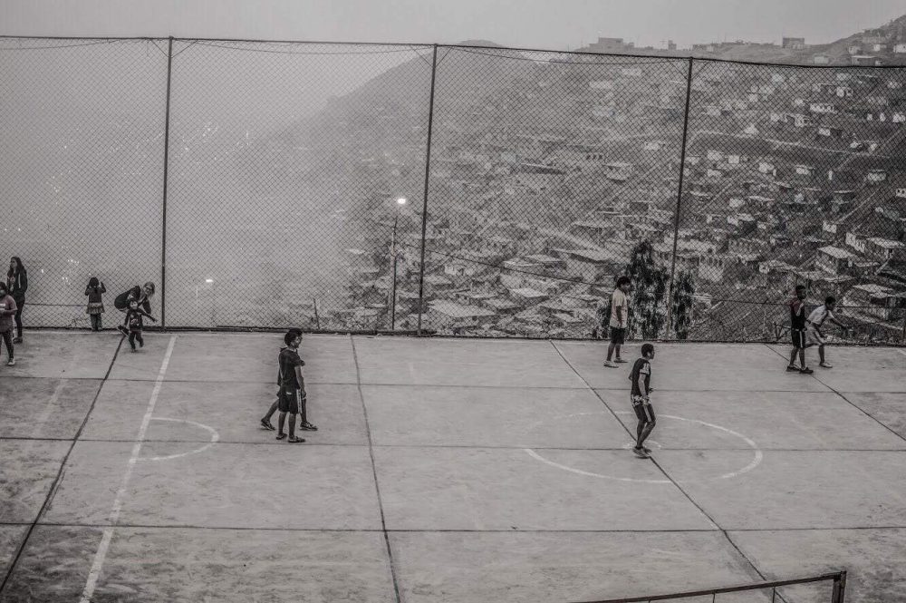A black and white photo of children playing soccer on a court in front of city buildings covering a hillside.