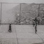 A black and white photo of children playing soccer on a court in front of city buildings covering a hillside.