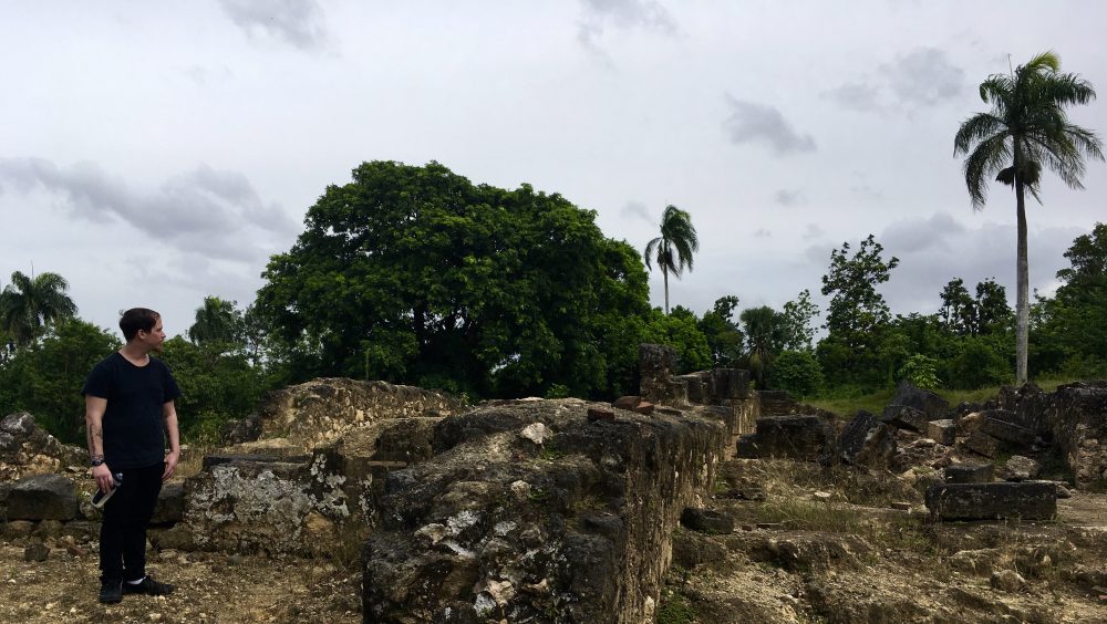 Photo of a student standing next to ruins with trees in the background.