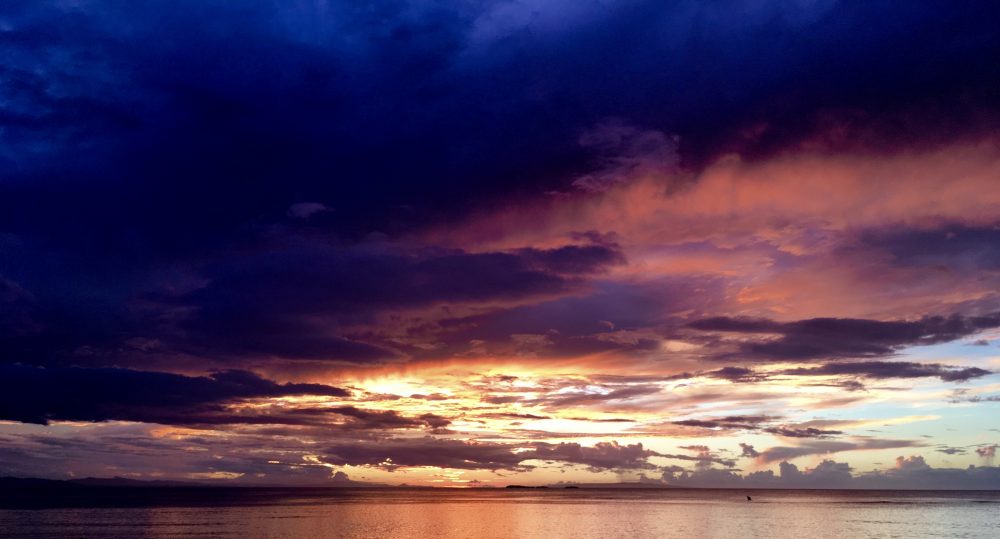A photo of a sunset with dark purple clouds on top, leading into yellow and orange clouds by the sea.