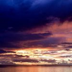 A photo of a sunset with dark purple clouds on top, leading into yellow and orange clouds by the sea.