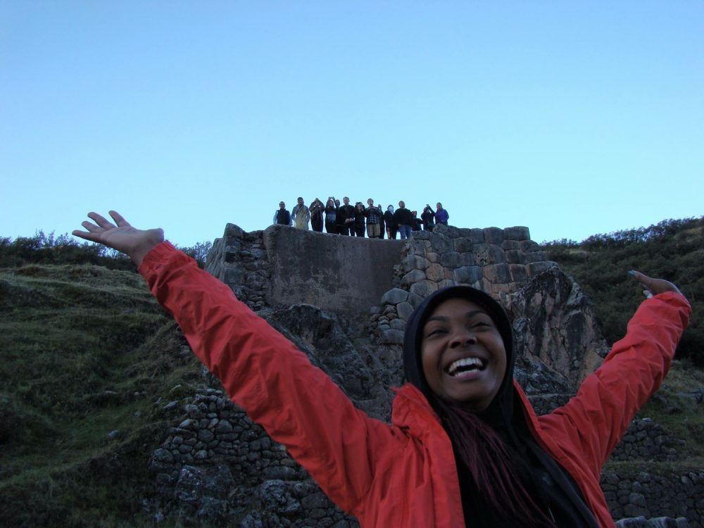 Photo of Sade Mitchell in front of a rock wall, with her fellow program participants standing on top.