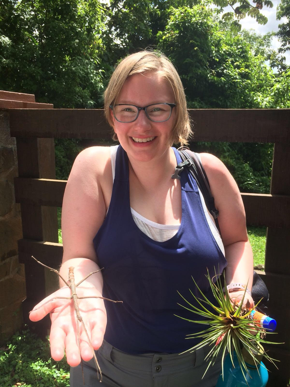 Hannah Hess, 2018-19 EAO Peer Mentor, blonde haired female wearing round blue glasses and a blue tank top holding a walking stick bug in her right hand and grass and an orange sunscreen bottle in her left hand, standing in front of a wooden fence on a lawn in front of trees in the background.