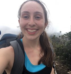 McKenna Erickson, a 2018-19 EAO peer mentor, is pictured wearing a gray backpack and blue and black tank top standing in the mountains of Costa Rica with dark green shrubs and thick gray fog in the background.