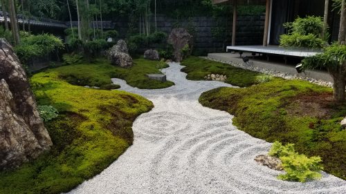 This photo shows a white rock garden in Japan. The rock garden is raked in a circular format and is shaped like a path between rocks and green moss on the left and right side. There is a small green plant coming out of the rock garden in the right foreground. Taller shrubs and trees are in the background.