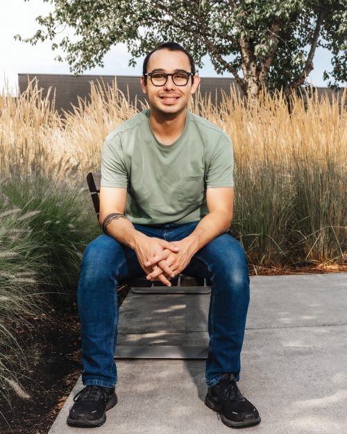 PCC student sitting on the edge of a bench with arms resting on knees and grass and trees behind him.