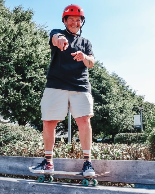 Skateboarder with red helmet, standing on top of skateboard deck and arms are outstretched and both pointer fingers are pointing ahead.