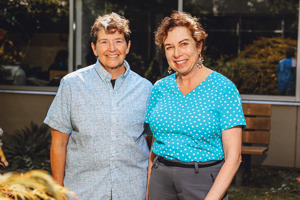 Person wearing a blue button up shirt and person wearing a turquoise shirt, standing next to each other in a landscaped area, smiling directly at the camera.