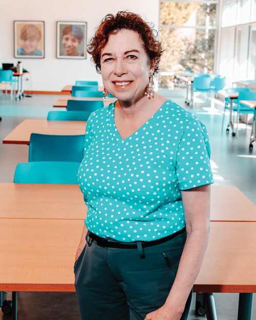 Person with turquoise shirt smiling at camera while standing in large room with tables and chairs.