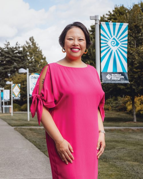 A woman wearing a pink dress standing on a path on PCC Rock Creek campus with lightpole flags in the background.