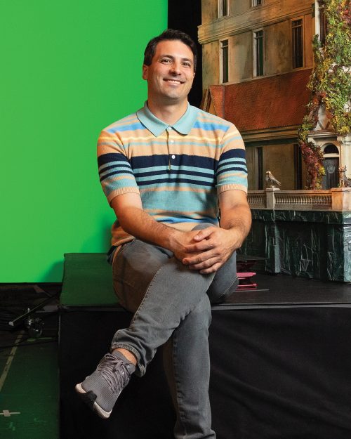 A person sitting on a stage set with left leg crossed over right leg and hands on left knee.