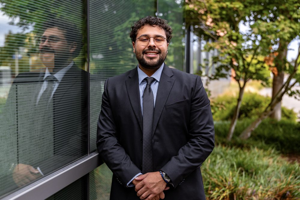 A man dressed in a suit and tie smiles at the camera while standing outdoors next to an exterior wall with vegetation in the background.