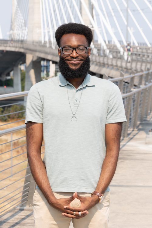 A man wearing glasses smiles at camera in front of Portland's Tilikum Bridge.
