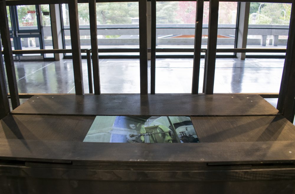 Black wooden box with video monitor in foreground, wooden rectilinear frame behind box, gallery windows in background.