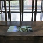 Black wooden box with video monitor in foreground, wooden rectilinear frame behind box, gallery windows in background.