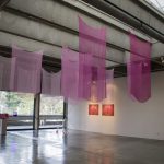 Art gallery with rectangles of sheer pink fabrics hanging from the ceiling at the center of the room; to the right are 3 paintings hanging on the wall; on the left are a video monitor and lights; in the far corner of the room is a plastic chair sculpture.