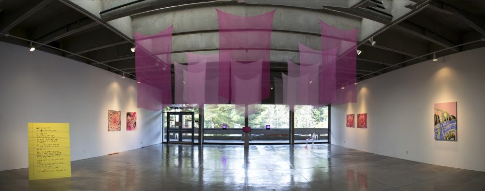 Panorama view of art gallery with sheer pink fabrics hanging from the ceiling in the center of the room; a large yellow board with writing in on the floor on the left; 2 pink paintings are on the left wall, 3 pink paintings are on the right wall; a pink video monitor and 2 pink lights are on the far wall in the center. In the background are large windows that reveal a view of the trees outside.