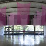Panorama view of art gallery with sheer pink fabrics hanging from the ceiling in the center of the room; a large yellow board with writing in on the floor on the left; 2 pink paintings are on the left wall, 3 pink paintings are on the right wall; a pink video monitor and 2 pink lights are on the far wall in the center. In the background are large windows that reveal a view of the trees outside.