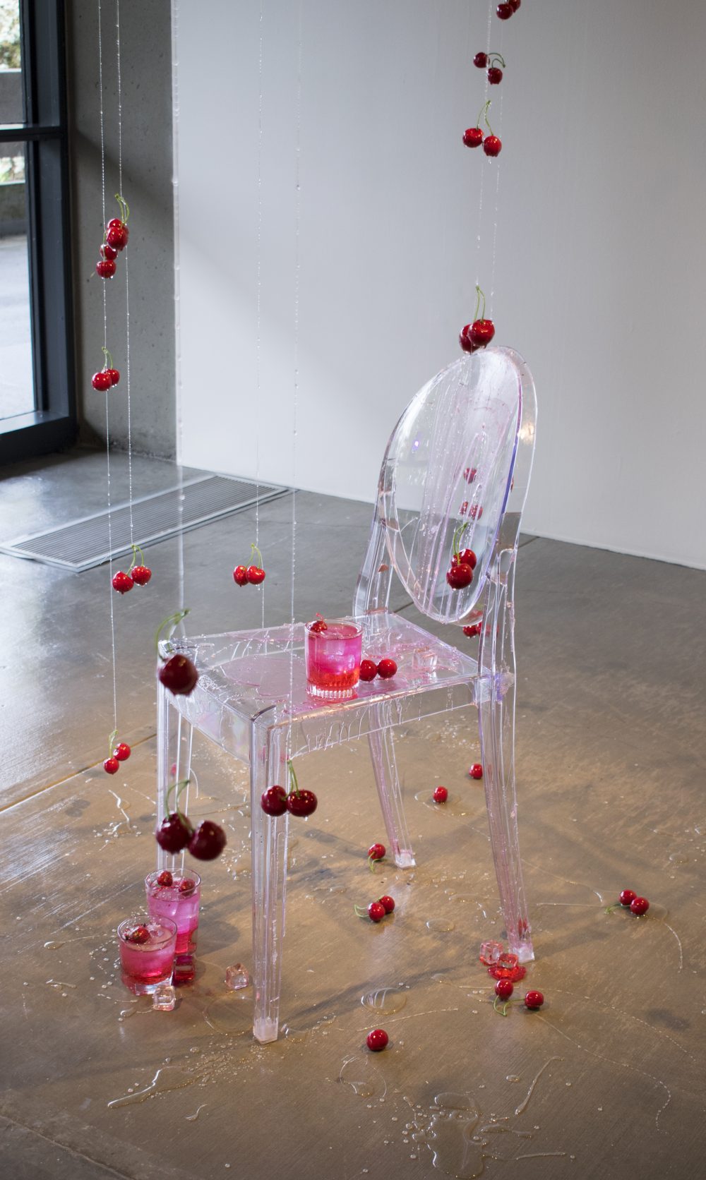 Detail of a larger sculpture; a clear plastic chair with a clear glass sitting on it; cherries are scattered on the floor and are hanging from the ceiling around and on top of the chair; there is a clear syrup dripped on the chair and floor.