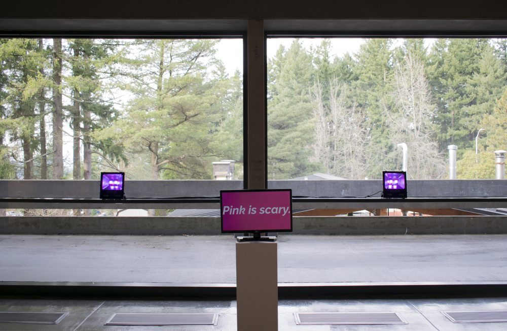 View of art installation in gallery with a video monitor in the center with the words "Pink is scary" on it and a pink light on either side of the monitor. Behind the installation are large windows that show a view of the trees outside.