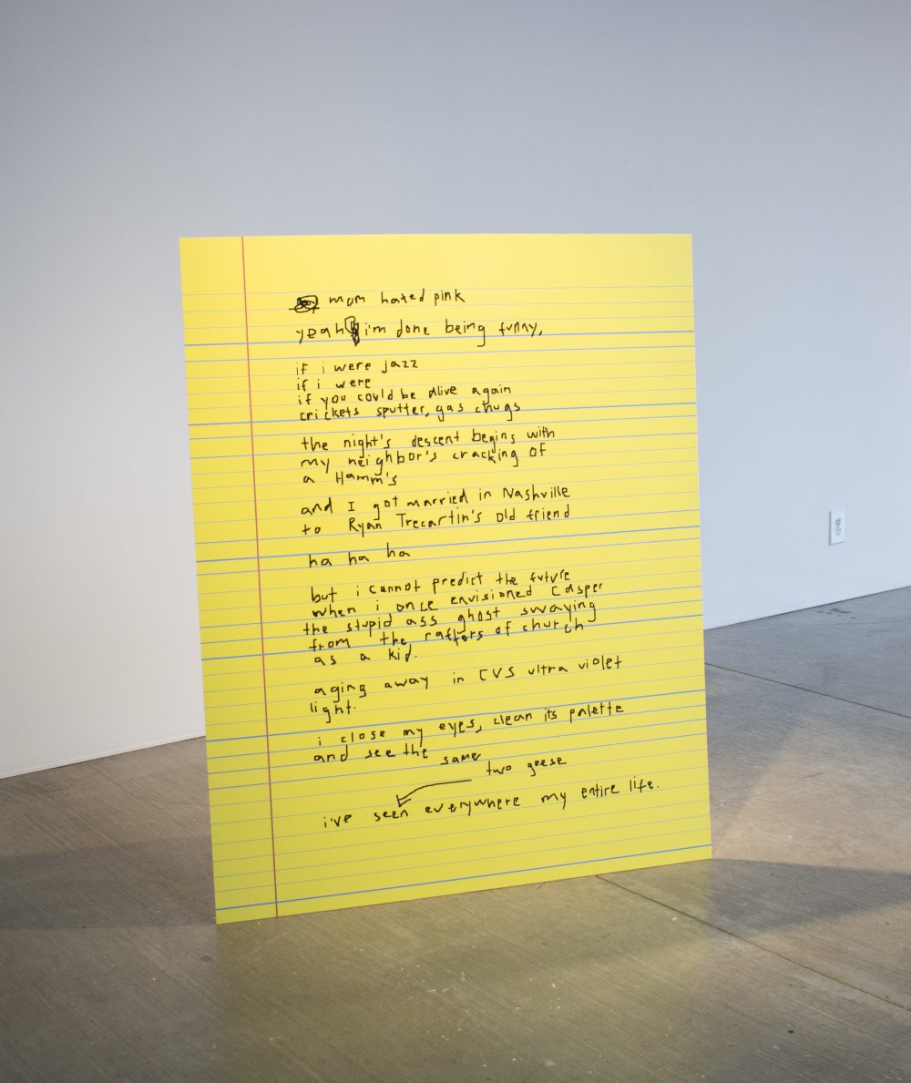Artwork made of a large yellow piece of cardboard that looks like a sheet from a legal pad, with handwritten text.