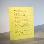 Artwork made of a large yellow piece of cardboard that looks like a sheet from a legal pad, with handwritten text.