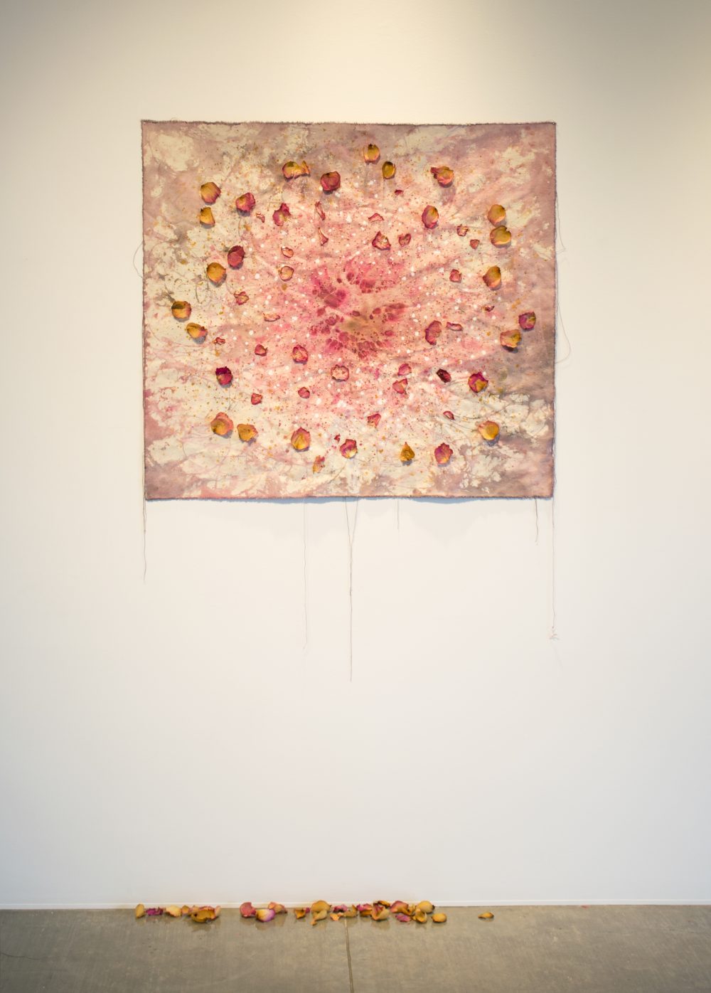 Artwork consisting of a piece of canvas that is painted pink and grey, with rose petals glued to it; this hangs on the wall and below are rose petals laying on the floor.