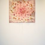Artwork consisting of a piece of canvas that is painted pink and grey, with rose petals glued to it; this hangs on the wall and below are rose petals laying on the floor.