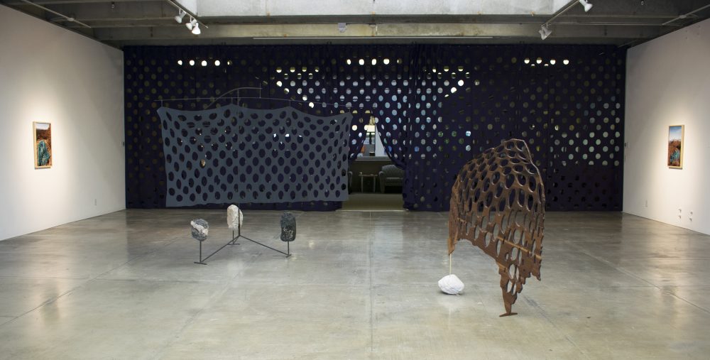 A sculpture on the left has 3 stones at its base and a grey screen with holes in it; a sculpture on the right has a wooden screen with holes in it standing on end with a stone as a base on one side. 2 landscape photos hang on either side; behind is a floor to ceiling, wall to wall dark curtain with a grid of large holes in it.