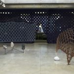 A sculpture on the left has 3 stones at its base and a grey screen with holes in it; a sculpture on the right has a wooden screen with holes in it standing on end with a stone as a base on one side. 2 landscape photos hang on either side; behind is a floor to ceiling, wall to wall dark curtain with a grid of large holes in it.