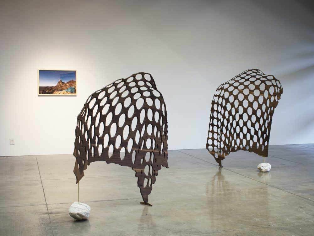 On the back wall is a landscape photo; on the gallery floor are two sculptures made of wood screens that have holes in them; each has a large white stone at its base.