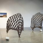On the back wall is a landscape photo; on the gallery floor are two sculptures made of wood screens that have holes in them; each has a large white stone at its base.