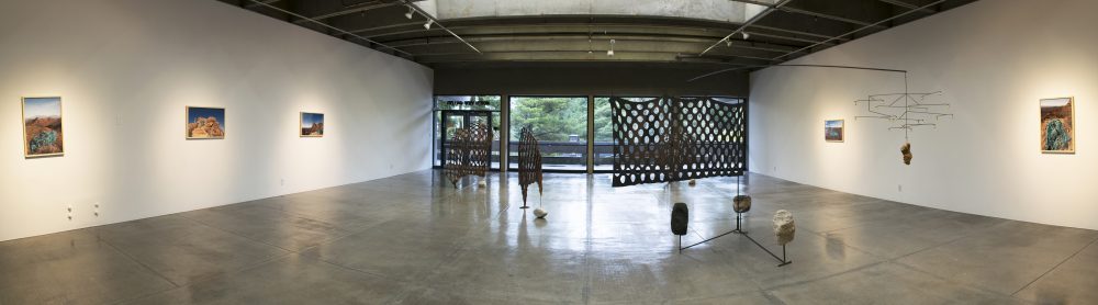 Panorama view of art gallery with landscape photos hanging on walls on the left and right sides and in the center stand 4 sculptures made of wood and stone and metal. In the background are floor to ceiling glass windows that show the trees outside.