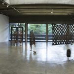 Panorama view of art gallery with landscape photos hanging on walls on the left and right sides and in the center stand 4 sculptures made of wood and stone and metal. In the background are floor to ceiling glass windows that show the trees outside.